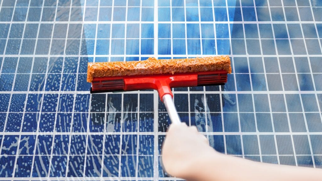 Close-up of a person's hand using a red squeegee with a brown absorbent sponge to clean a blue solar panel. The panel has a grid-like pattern with visible water streaks indicating the cleaning process, symbolizing maintenance for optimal solar energy production.