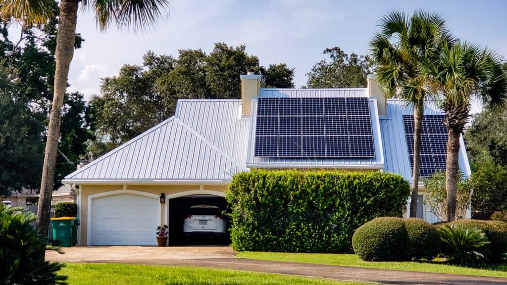 A modern suburban home with a white metal roof featuring multiple solar panels installed on the upper portion. The house has a two-car garage, a neatly trimmed hedge, and a well-maintained lawn with palm trees. A white car is partially visible inside the garage, and a green trash bin is placed near the driveway. The background consists of lush greenery and a partly cloudy sky.