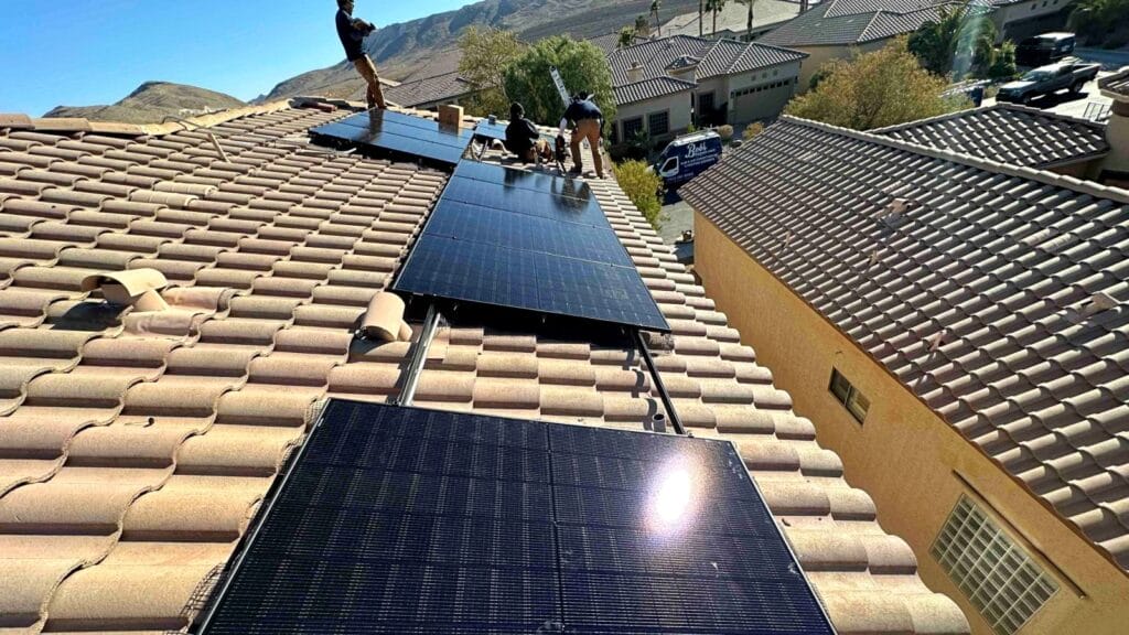 A team of workers from Bob's Repair installs black solar panels on a terracotta-tiled residential roof in a sunny neighborhood, with mountains and palm trees in the background.