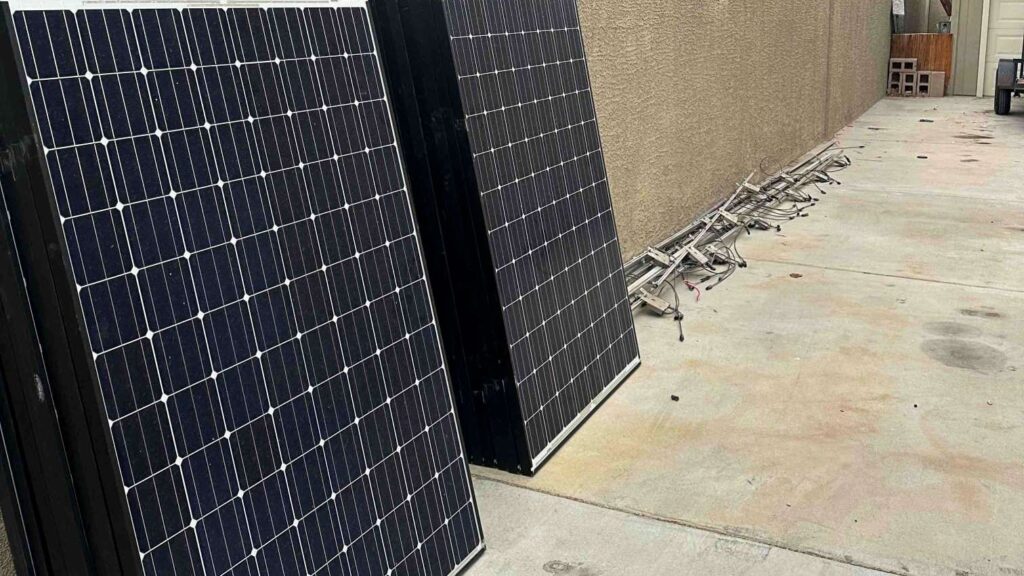 Stack of removed solar modules leaning against a yard wall, with mounting racks laid out nearby.