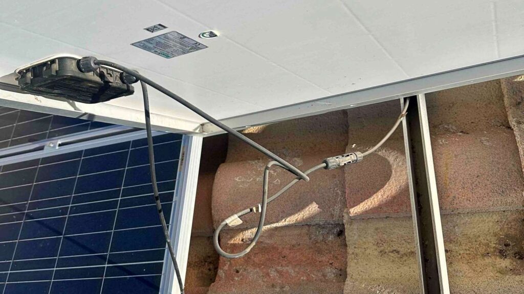 Underside of a solar panel showing the junction box and DC wiring connections above roof tiles.