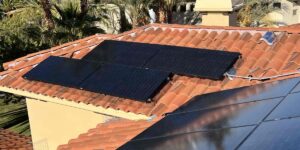 Two black solar panel arrays are mounted on a red clay tile roof next to a yellow stucco chimney, with palm trees and another home visible in the background of this Las Vegas neighborhood, illustrating how solar panel efficiency changes at 115 degrees and where solar panel heat loss occurs.